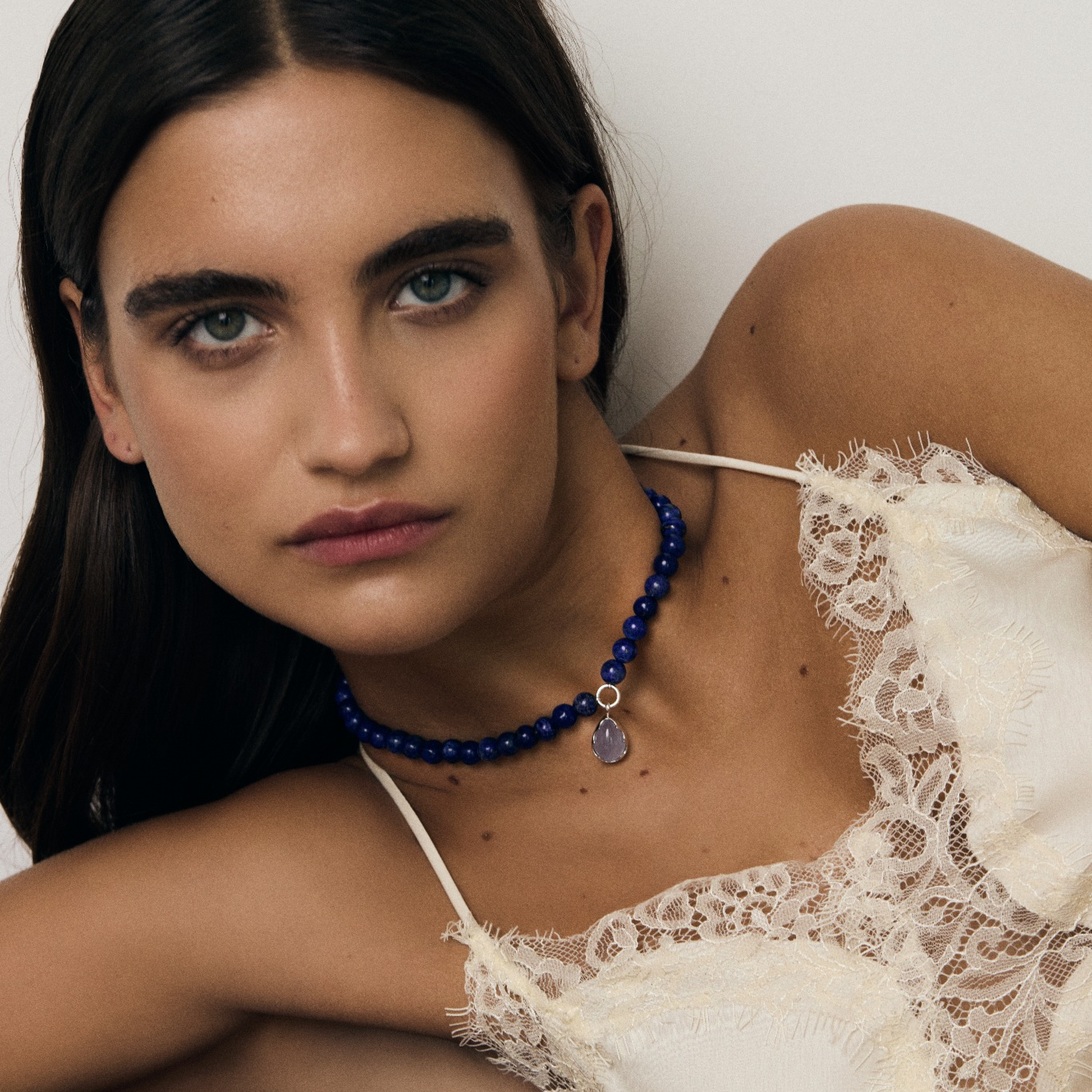 Woman wearing a white lace top and blue necklace on a plain background