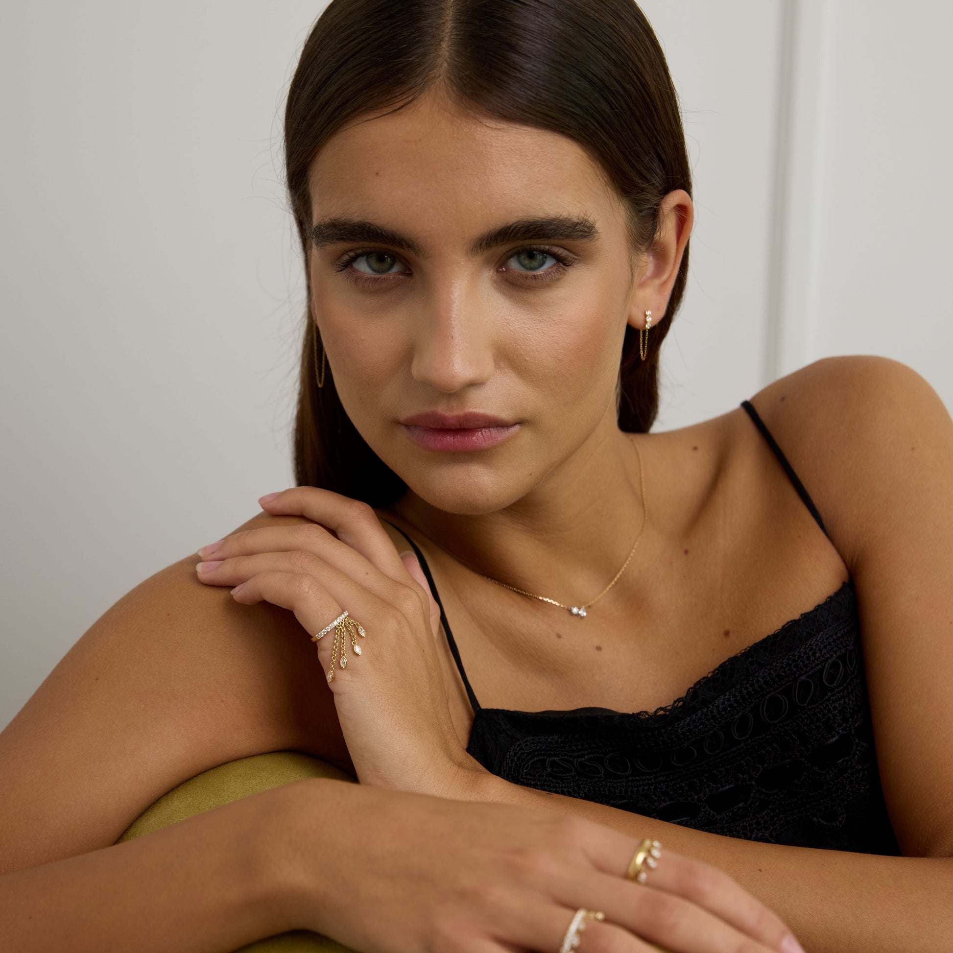 Woman wearing gold jewelry sitting on a green cushion against a white wall