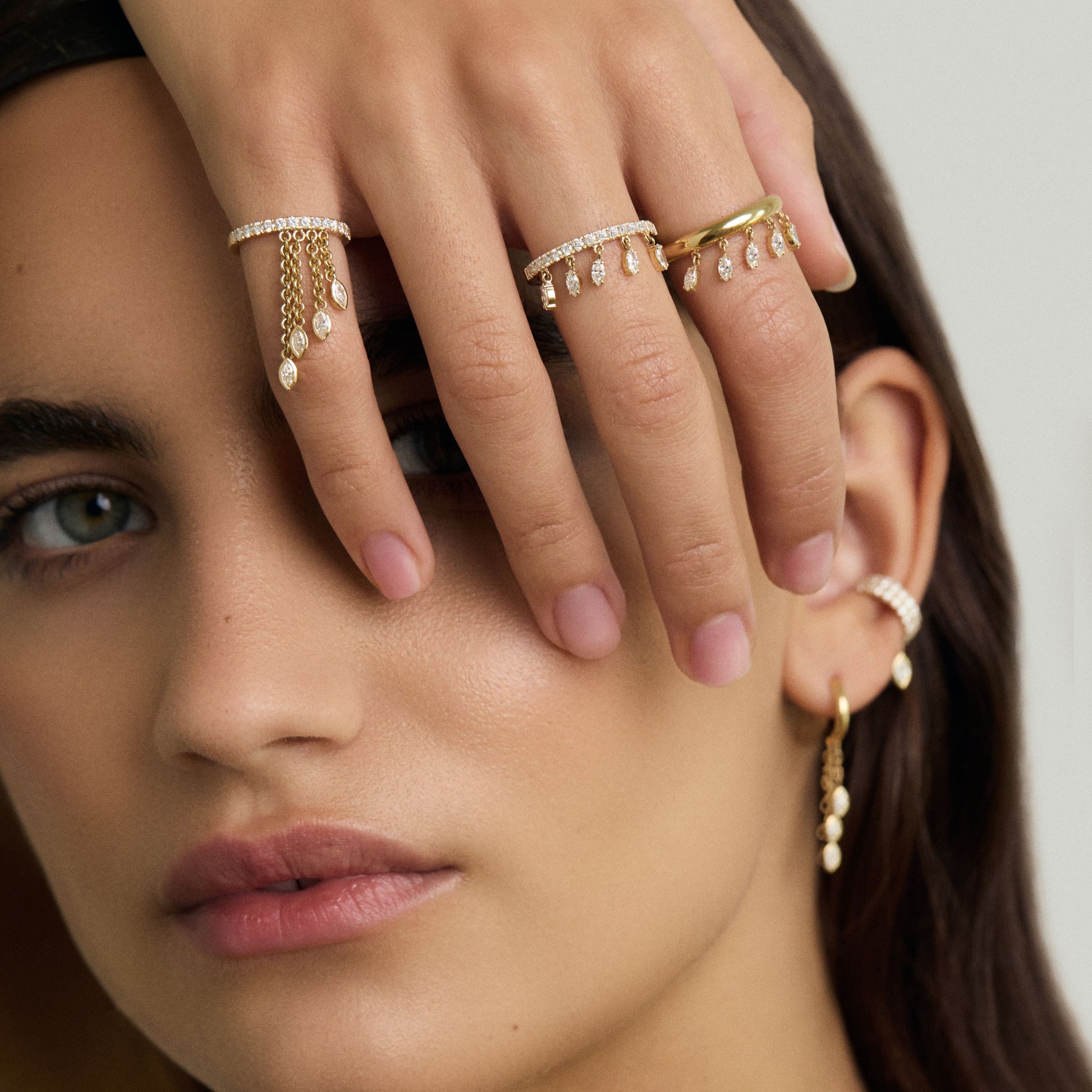 Close-up of a woman's face with gold jewelry on her fingers and ear.